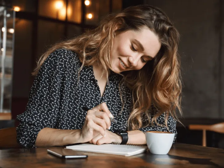 A woman creatively inspired, writing in a notebook, fueled by a cup of coffee.