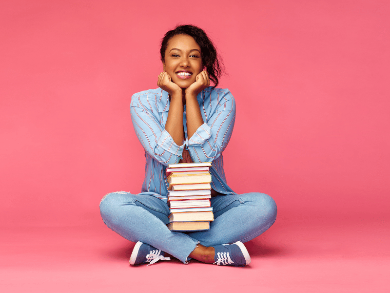 A young black woman surrounded by a stack of books, perfect for gifts for writers.