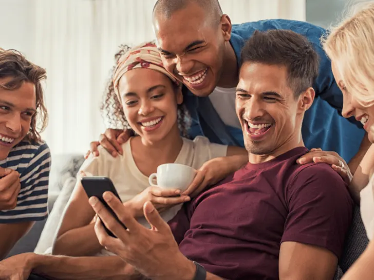 A group of friends laughing while using a cell phone and sharing their favorite catch phrase.