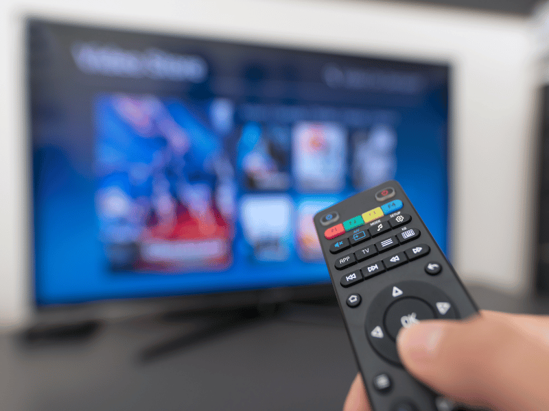 A person holding a remote control in front of a TV, trying to decide between different video streaming services.